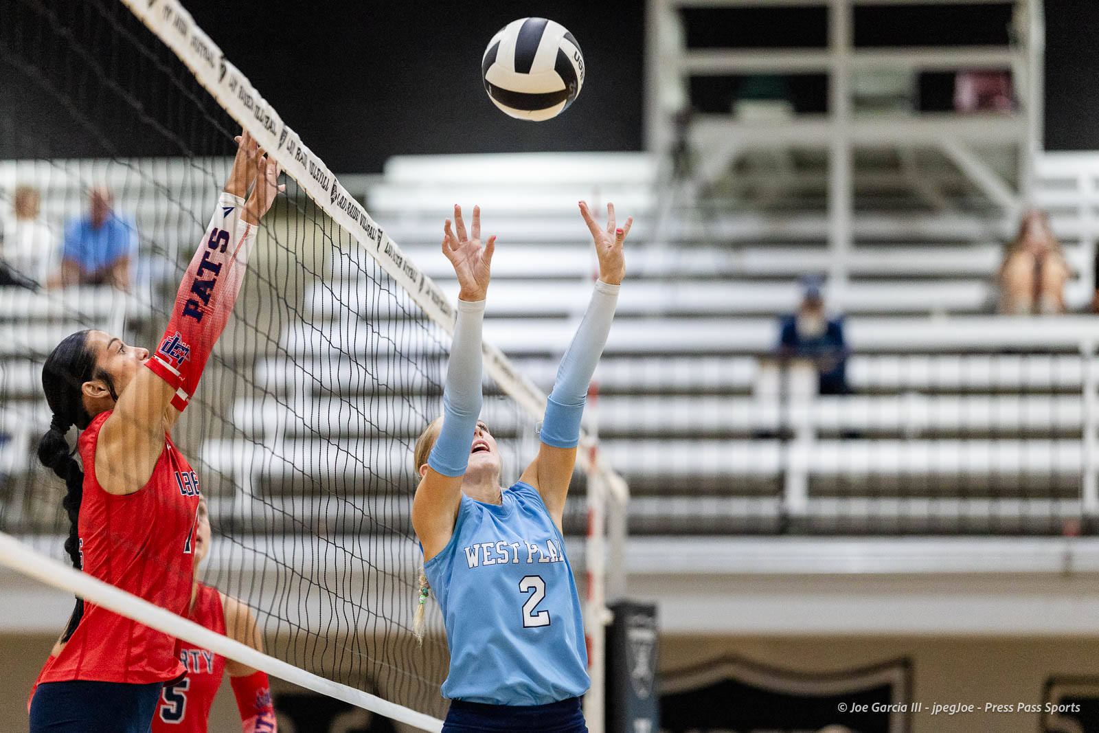 randall-showcase-2026-jpegJoe-pps-22 | Press Pass Sports Volleyball players reach to block the ball at the net during a match, one in red and one in blue jerseys, gym bleachers in the background.