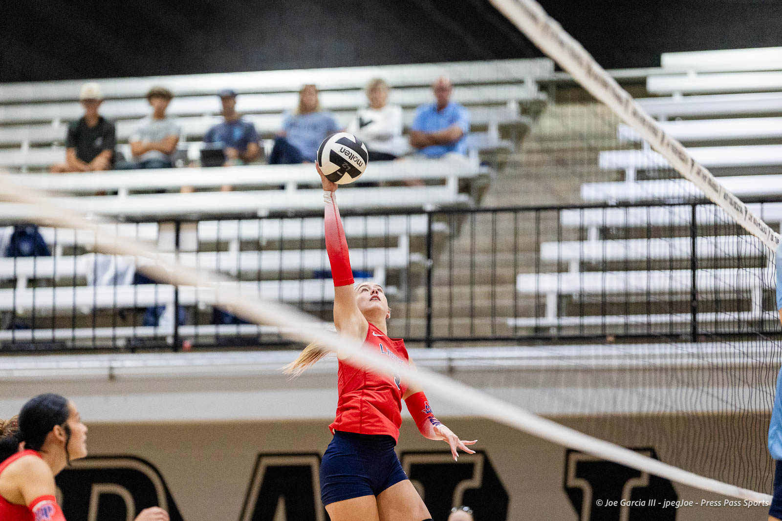 randall-showcase-2026-jpegJoe-pps-24 | Press Pass Sports Female volleyball player in a red jersey jumps to spike the ball over the net in an indoor gym, with spectators in the stands behind him/her.
