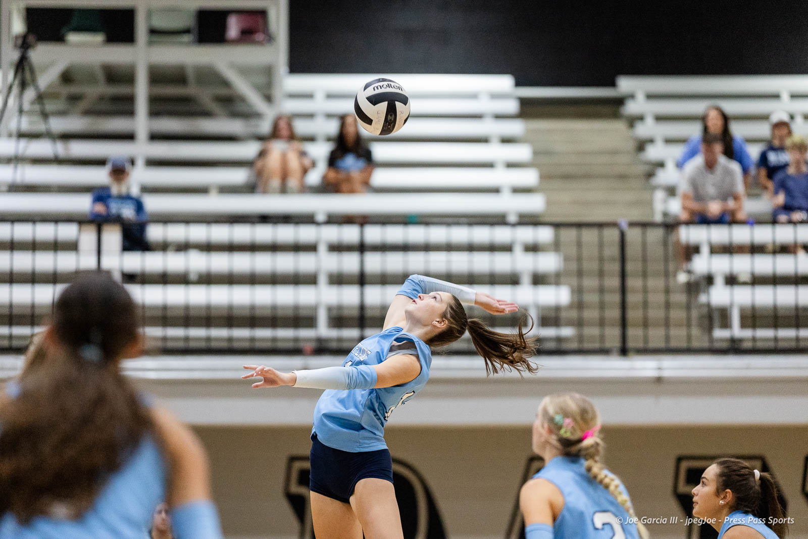 randall-showcase-2026-jpegJoe-pps-25 | Press Pass Sports Female volleyball player in light blue jersey serving or spiking the ball during a match in a gym, ball in midair with spectators in bleachers behind.