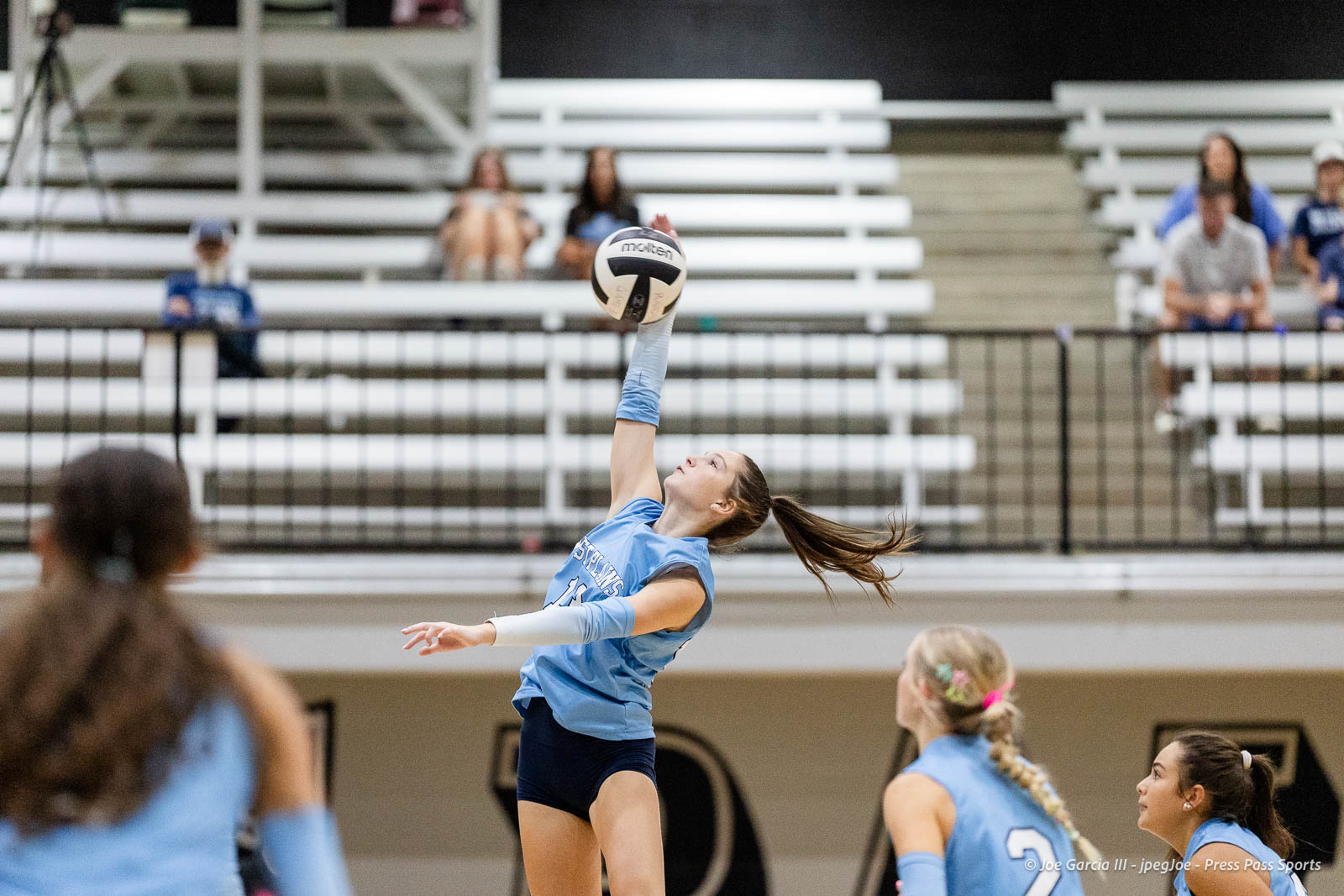 randall-showcase-2026-jpegJoe-pps-26 | Press Pass Sports Female volleyball player in a light blue jersey jumps to spike the ball during an indoor match, with spectators in white bleachers in the background.