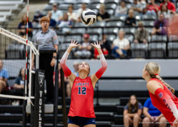 randall-showcase-2026-jpegJoe-pps-32 | Press Pass Sports Liberty player #12 in red uniform raises hands to set the volleyball during a rally, with a referee and audience in the background.