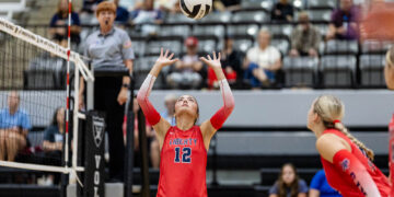 Liberty player #12 in red uniform raises hands to set the volleyball during a rally, with a referee and audience in the background.