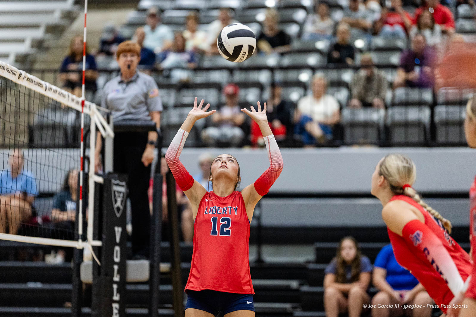 randall-showcase-2026-jpegJoe-pps-32 | Press Pass Sports Liberty player #12 in red uniform raises hands to set the volleyball during a rally, with a referee and audience in the background.