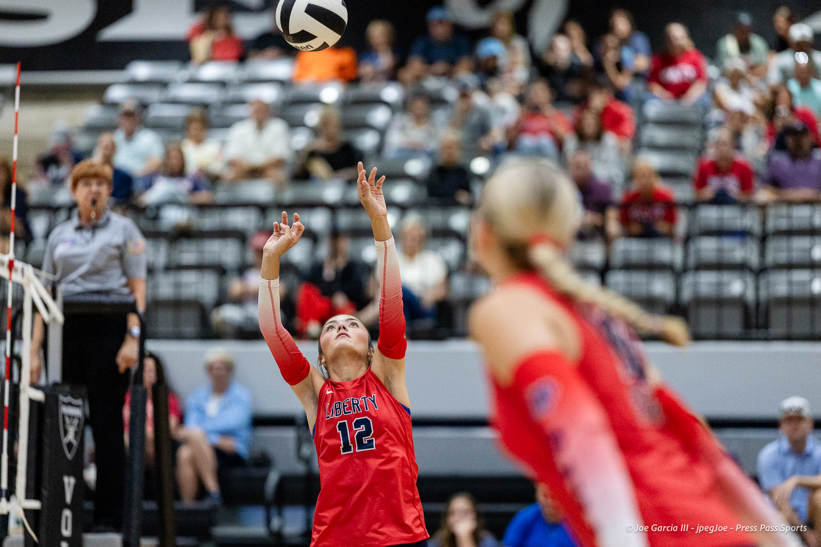 randall-showcase-2026-jpegJoe-pps-34 | Press Pass Sports Female volleyball player in a red jersey (number 12) reaching up to set the ball during a match with spectators in the stands behind her.