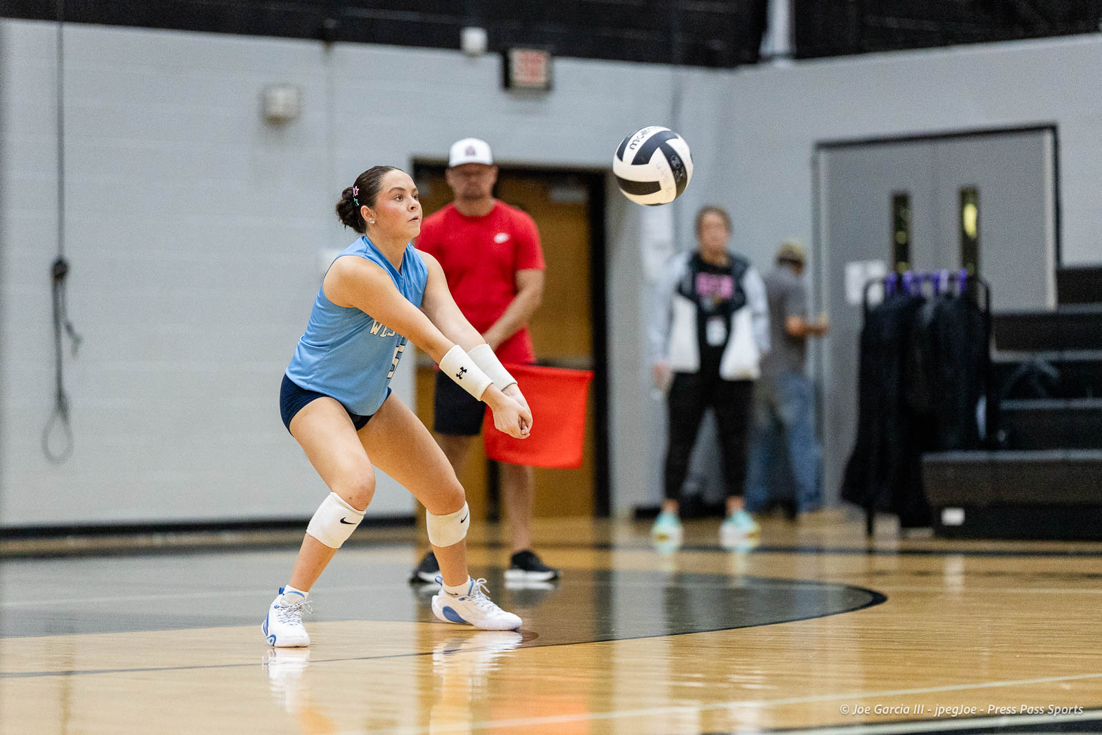 randall-showcase-2026-jpegJoe-pps-35 | Press Pass Sports Female volleyball player in a light blue jersey and knee pads bends forward to dig while a ball approaches in a gym court.
