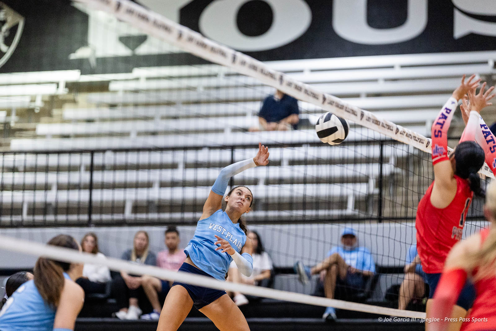 randall-showcase-2026-jpegJoe-pps-36 | Press Pass Sports Female volleyball player in a light-blue jersey spikes the ball over the net as red-jersey blockers jump to defend on the other side, in a gym with spectators in the background.