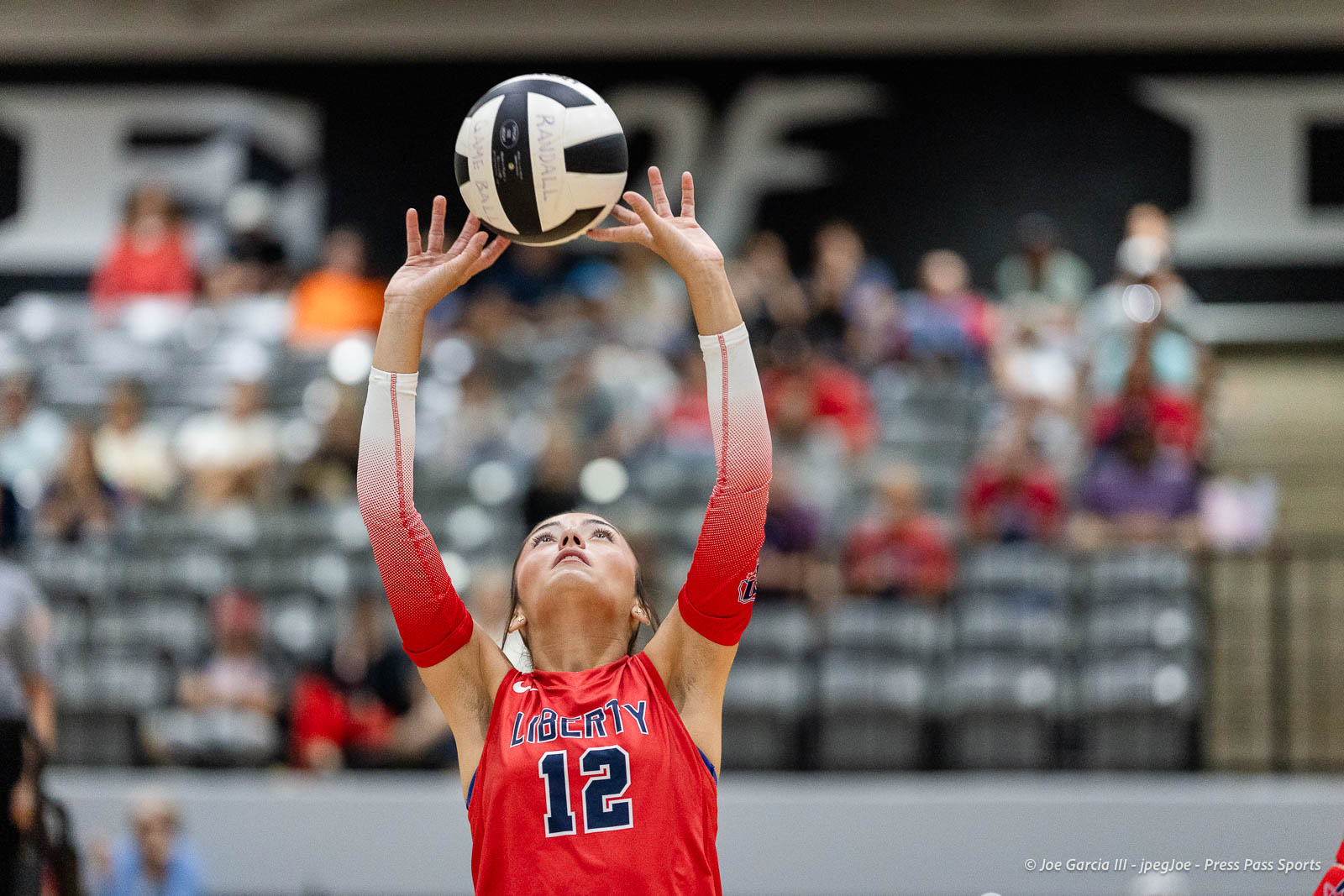 randall-showcase-2026-jpegJoe-pps-38 | Press Pass Sports Volleyball player in red Liberty jersey #12 sets a ball above her head during a match with blurred spectators in the stands behind her.