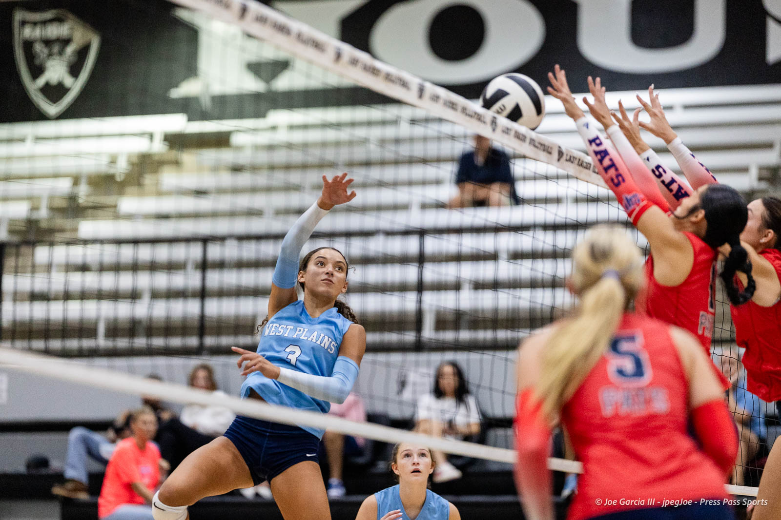 randall-showcase-2026-jpegJoe-pps-41 | Press Pass Sports Female volleyball player in a blue West Plains jersey (number 3) jumps to spike the ball over the net as opponents in red jerseys reach to block.