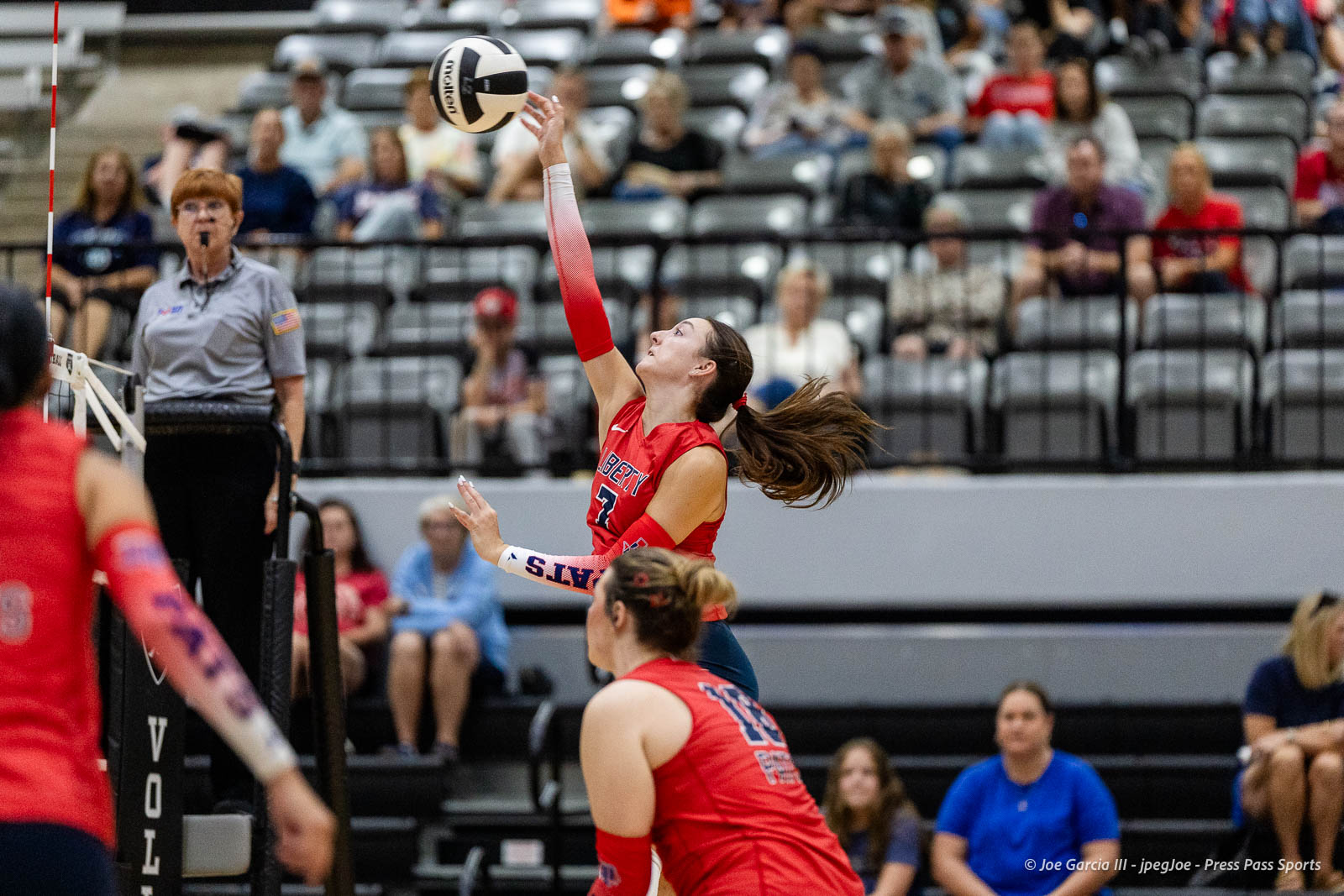 randall-showcase-2026-jpegJoe-pps-43 | Press Pass Sports Female volleyball player in a red uniform jumping to contact the ball over the net, with a referee watching near the net and spectators in the stands behind her.