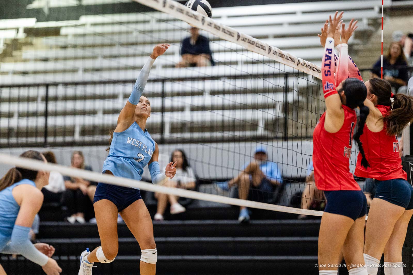 randall-showcase-2026-jpegJoe-pps-44 | Press Pass Sports Female volleyball player in a light blue West Plains jersey jumps to spike the ball over the net as red-team blockers reach up in defense.