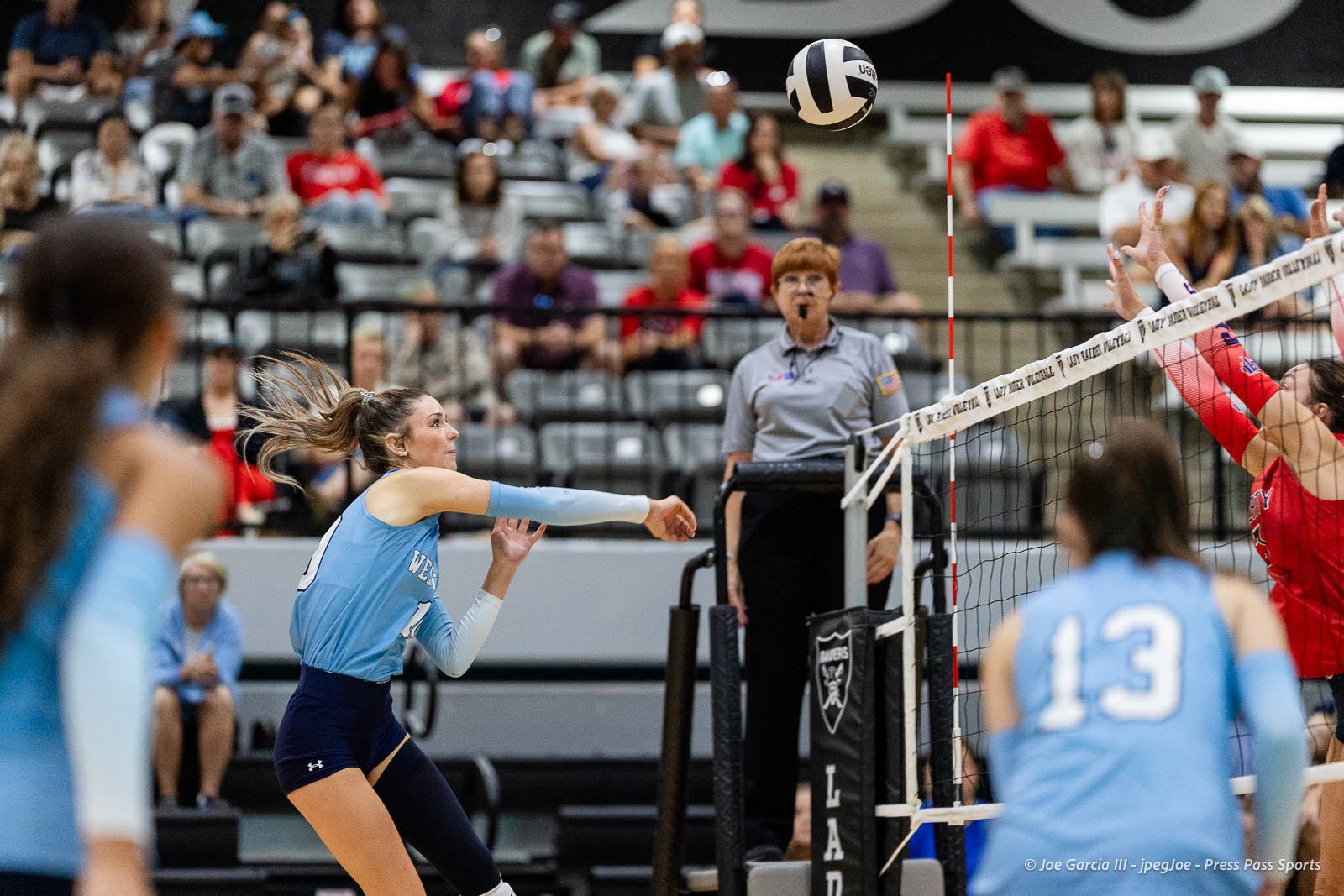 randall-showcase-2026-jpegJoe-pps-45 | Press Pass Sports Female volleyball player in light blue jersey hits ball over the net during a match in an indoor arena with spectators in the stands.