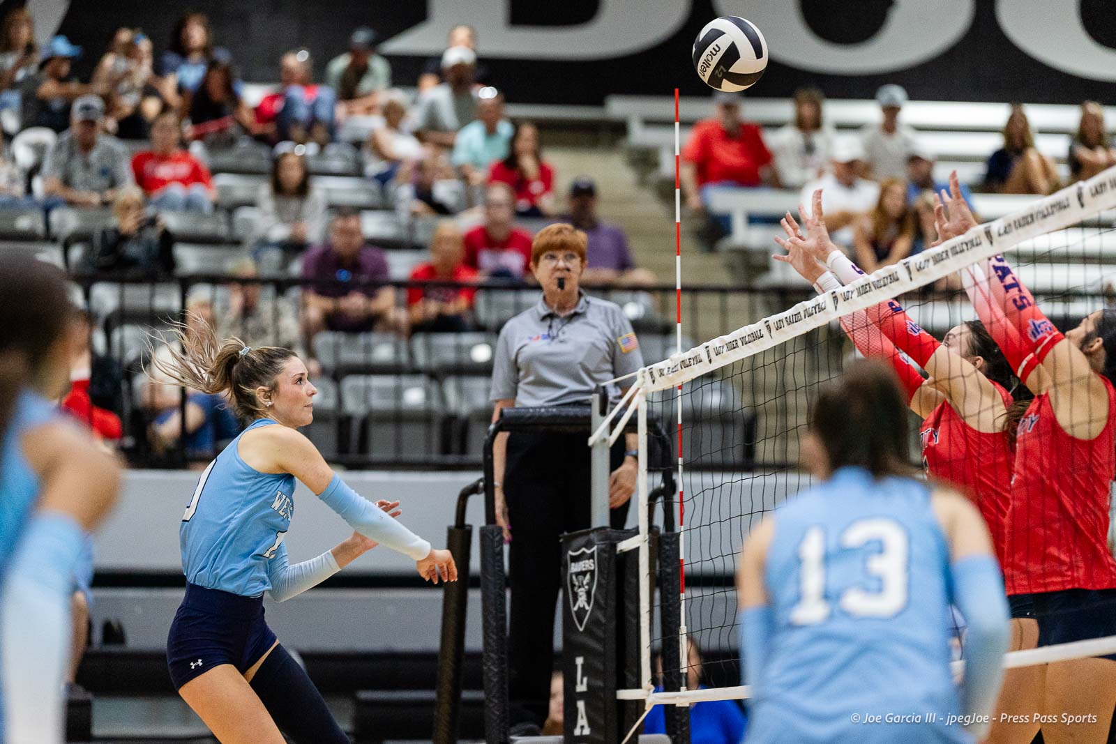 randall-showcase-2026-jpegJoe-pps-46 | Press Pass Sports Action at a women's indoor volleyball match: a blue-team player in motion near the net as the ball sails above, while red-team players jump to block; referee stands in the background watching, with spectators in the stands.