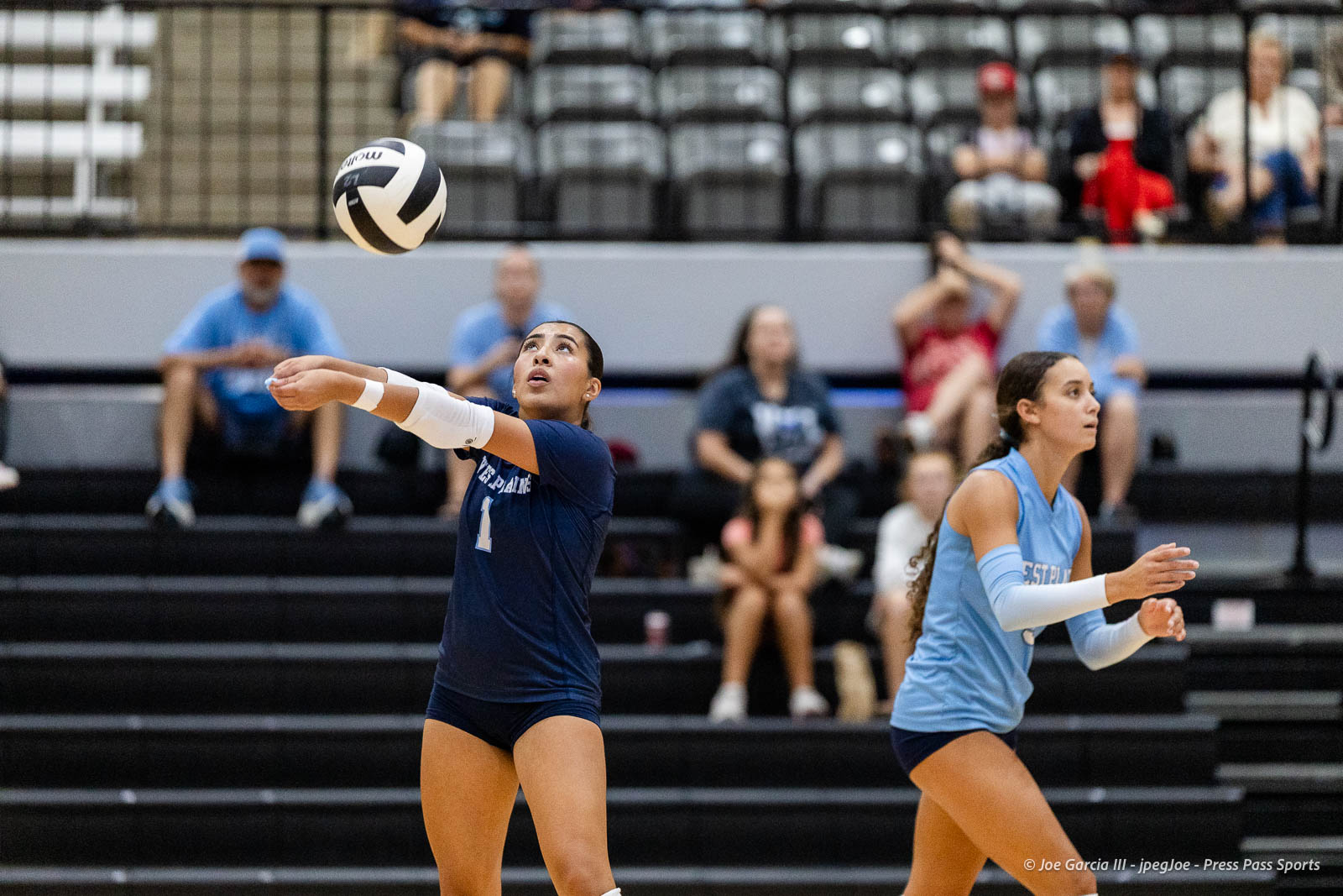 randall-showcase-2026-jpegJoe-pps-47 | Press Pass Sports Two female volleyball players—one in navy, one in light blue—prepare for a return as the ball flies overhead during a match, with spectators in the stands blurred behind them.