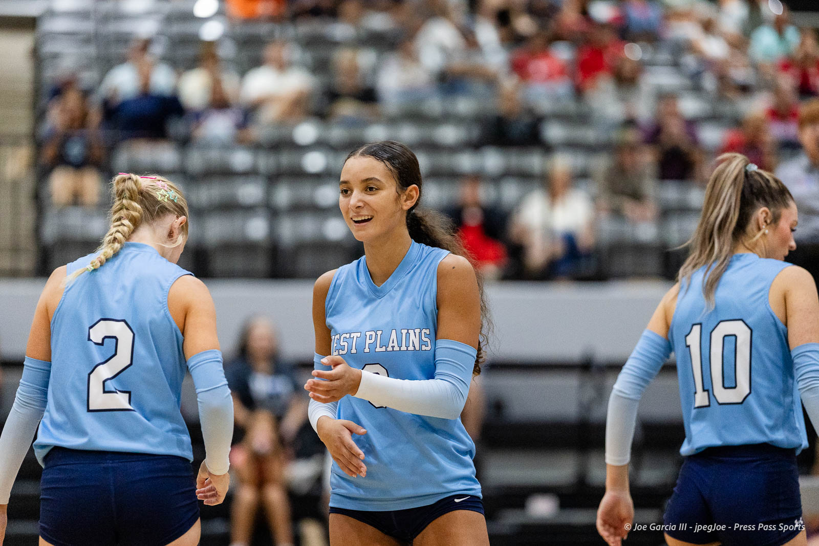 randall-showcase-2026-jpegJoe-pps-48 | Press Pass Sports Three female volleyball players in light blue jerseys on a gym court, smiling and talking between plays, with numbers 2 and 10 visible on their backs and a blurred spectator crowd in the stands.