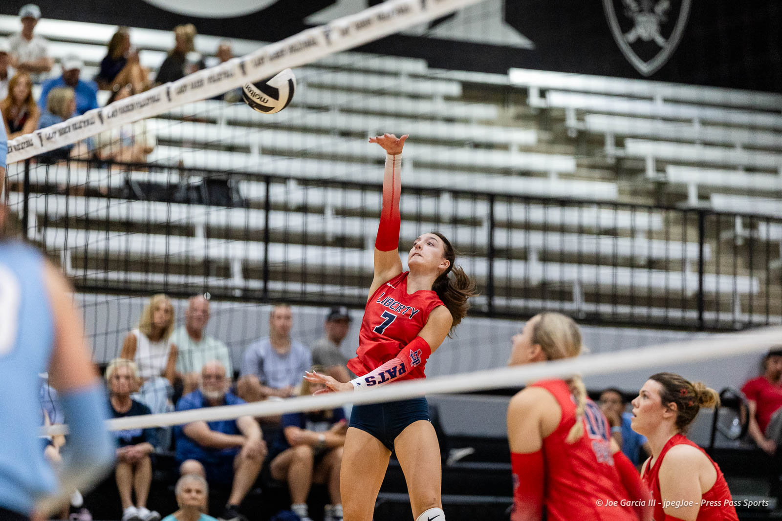 randall-showcase-2026-jpegJoe-pps-50 | Press Pass Sports Female volleyball player in red jersey leaps to spike the ball over the net during a match, with fans in the bleachers.