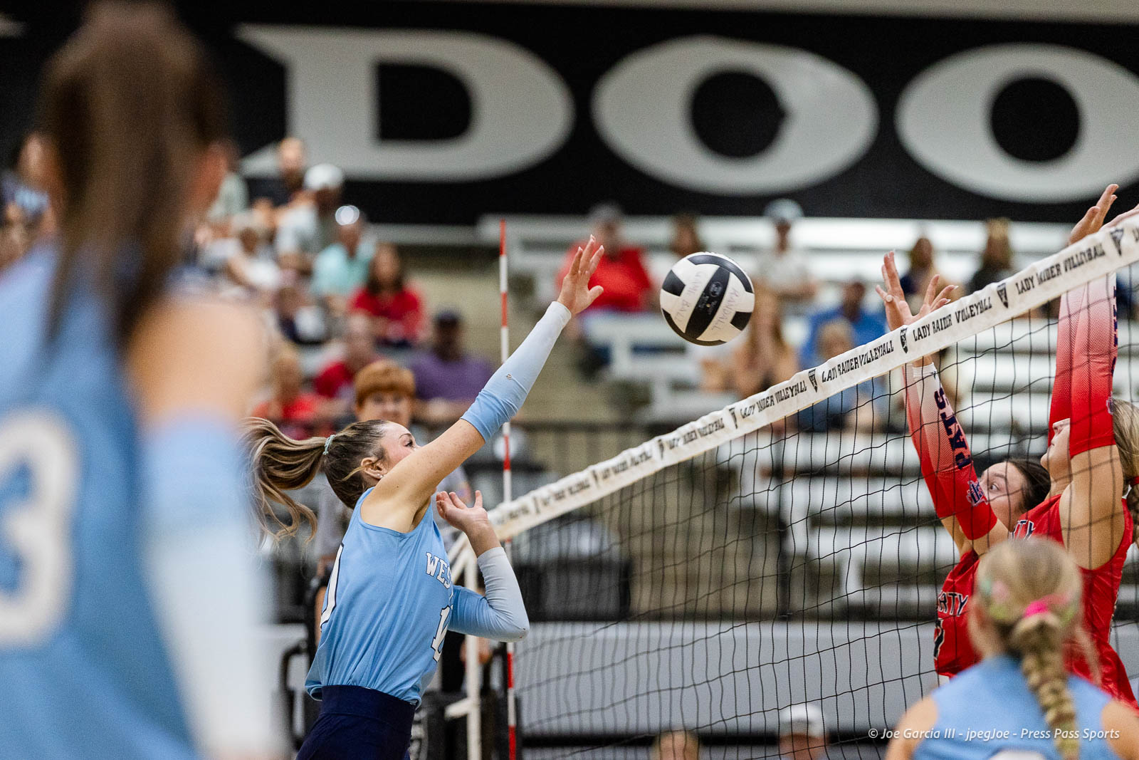 randall-showcase-2026-jpegJoe-pps-51 | Press Pass Sports Blue-uniform volleyball player spikes the ball over the net toward red-team blockers with a crowded gym in the background.