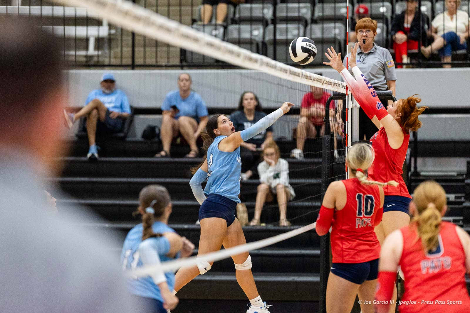 randall-showcase-2026-jpegJoe-pps-53 | Press Pass Sports Volleyball action at the net: blue team player spikes the ball while red team blocks, spectators in background and a referee nearby.