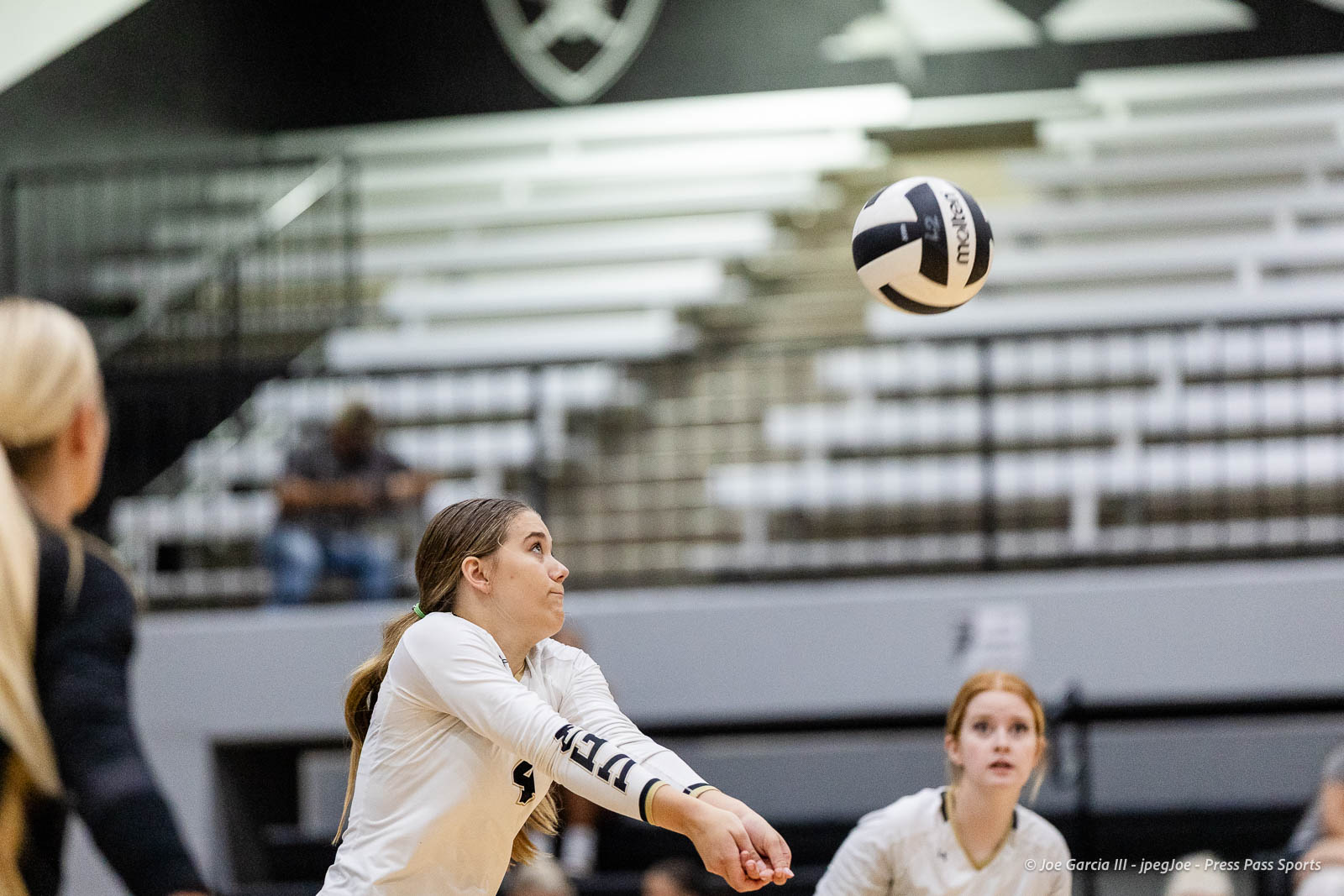 randall-showcase-2026-jpegJoe-pps-56 | Press Pass Sports Female volleyball player in a white jersey extends arms to receive the ball in an indoor gym, with teammates and bleachers in the background.
