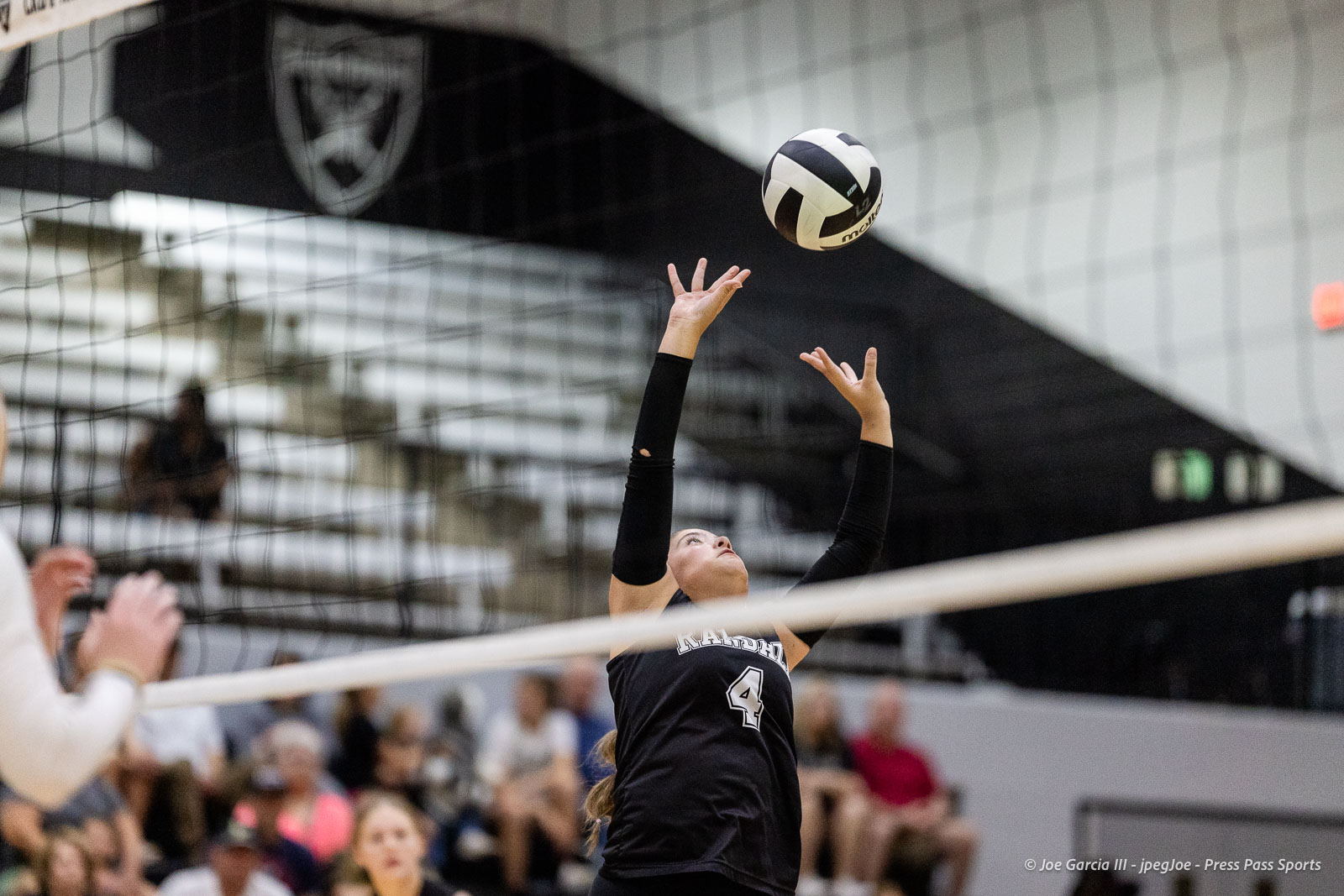 randall-showcase-2026-jpegJoe-pps-57 | Press Pass Sports Female volleyball player in a black uniform (#4) reaches up to set the ball over the net during a match.