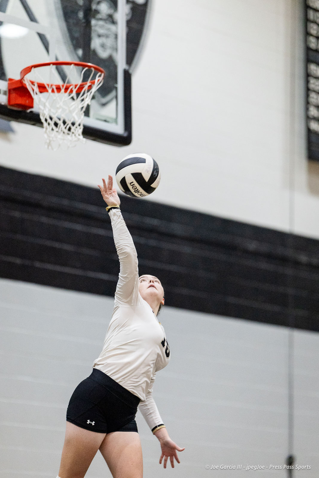 randall-showcase-2026-jpegJoe-pps-58 | Press Pass Sports Female volleyball player in a beige long-sleeve shirt and black shorts reaches up to spike a ball indoors near a basketball hoop in a gym.