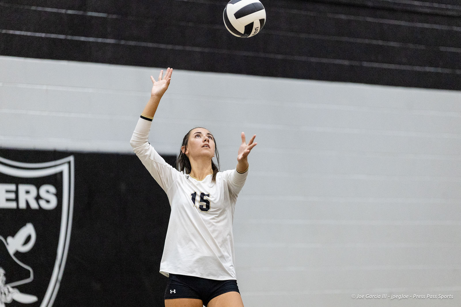 randall-showcase-2026-jpegJoe-pps-59 | Press Pass Sports Female volleyball player in a white jersey with the number 15, reaching up to set the ball in an indoor gym.