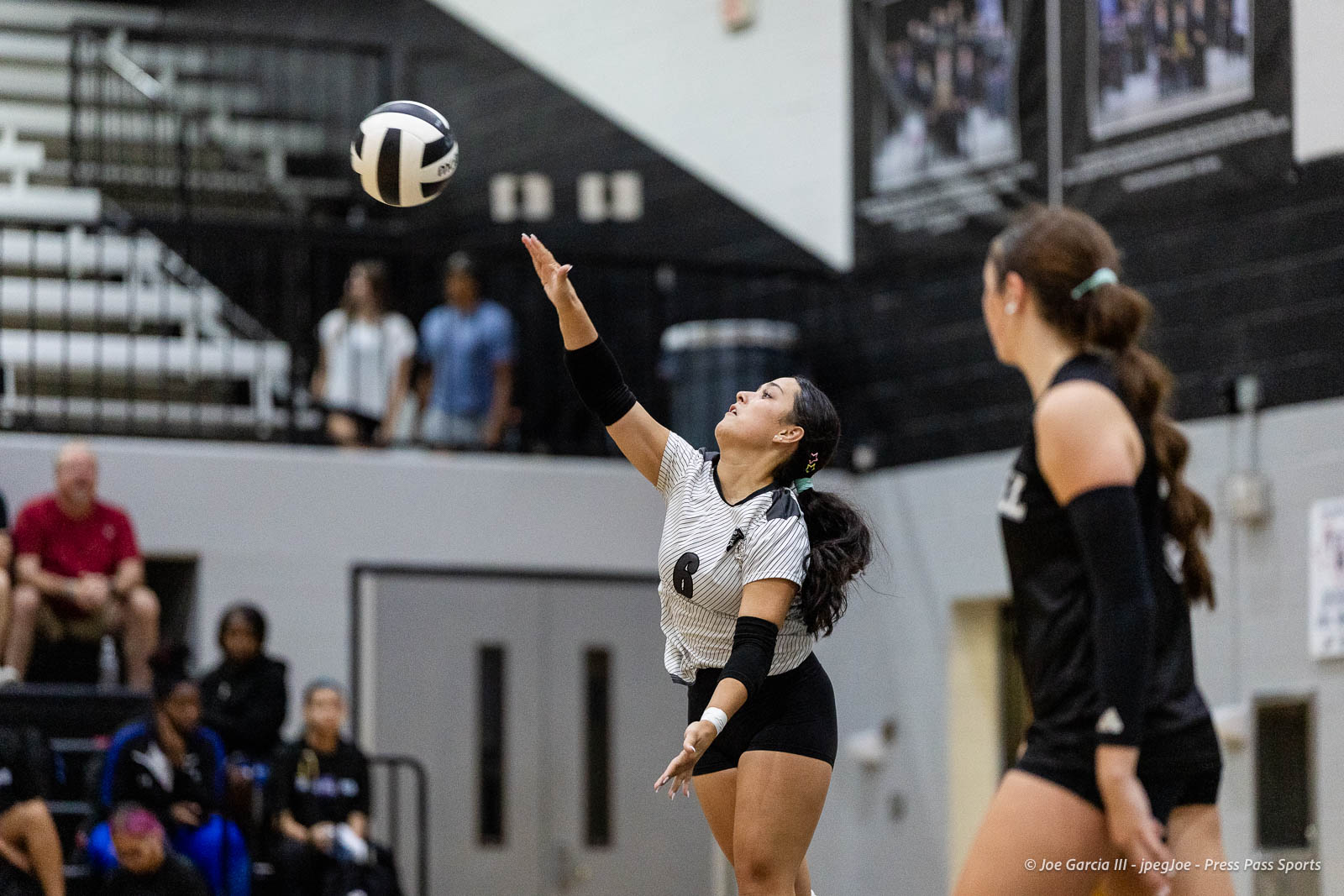 randall-showcase-2026-jpegJoe-pps-62 | Press Pass Sports Female volleyball player in a striped jersey serves the ball in a gym, with teammates and spectators in the background.
