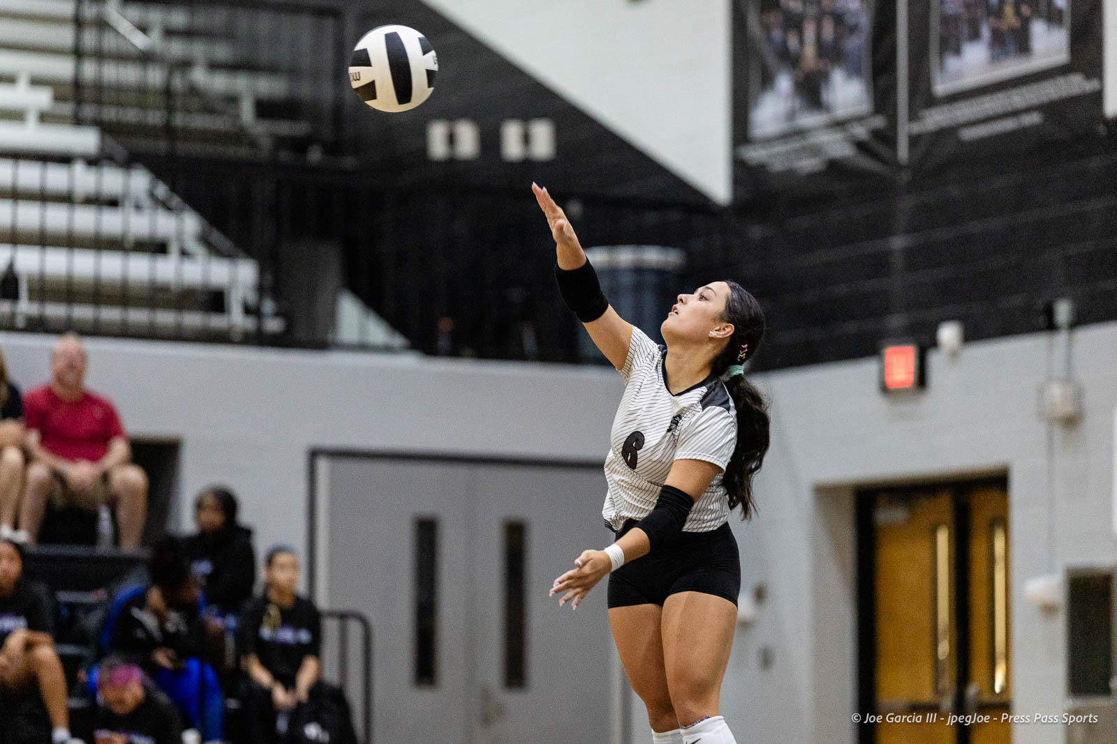 randall-showcase-2026-jpegJoe-pps-64 | Press Pass Sports Female volleyball player in a black-and-white striped jersey serves the ball in an indoor gym, with spectators in the stands behind her.