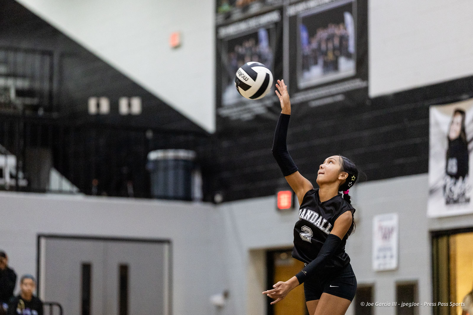randall-showcase-2026-jpegJoe-pps-65 | Press Pass Sports Female volleyball player in a black uniform serves the ball indoors on a gym court, reaching upward with extended arm.