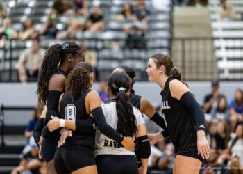 randall-showcase-2026-jpegJoe-pps-67 | Press Pass Sports Volleyball players in black uniforms form a huddle on the court with arms around each other, fans in bleachers behind them.