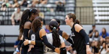 Volleyball players in black uniforms form a huddle on the court with arms around each other, fans in bleachers behind them.