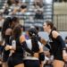 Volleyball players in black uniforms form a huddle on the court with arms around each other, fans in bleachers behind them.