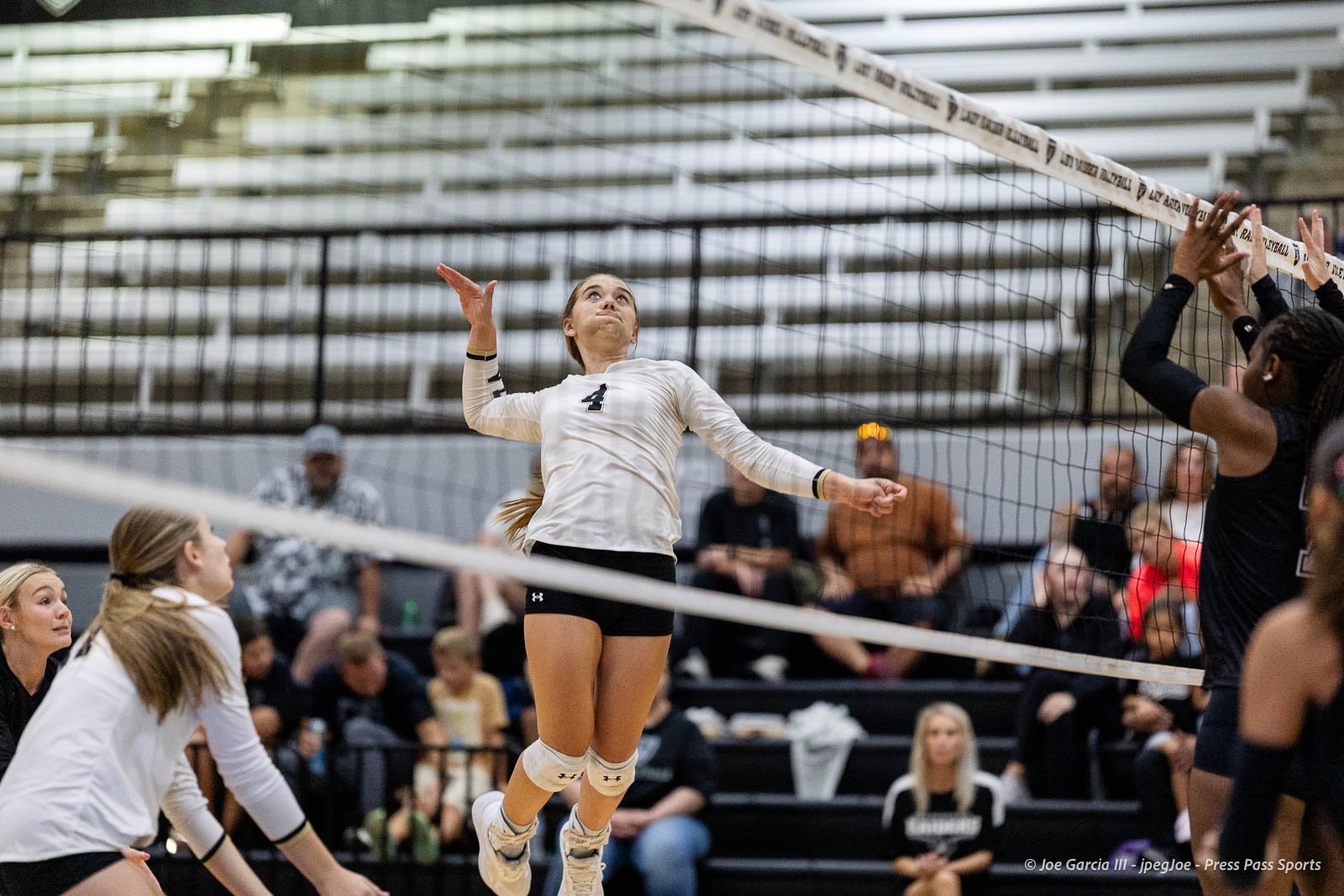 randall-showcase-2026-jpegJoe-pps-69 | Press Pass Sports Female volleyball player in a white jersey jumps for a spike near the net, with teammates and spectators in the bleachers behind.
