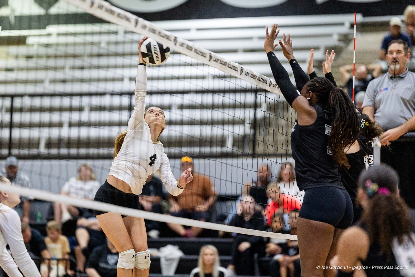 randall-showcase-2026-jpegJoe-pps-70 | Press Pass Sports Female volleyball player in white jumps to spike the ball over the net as opponents block nearby.