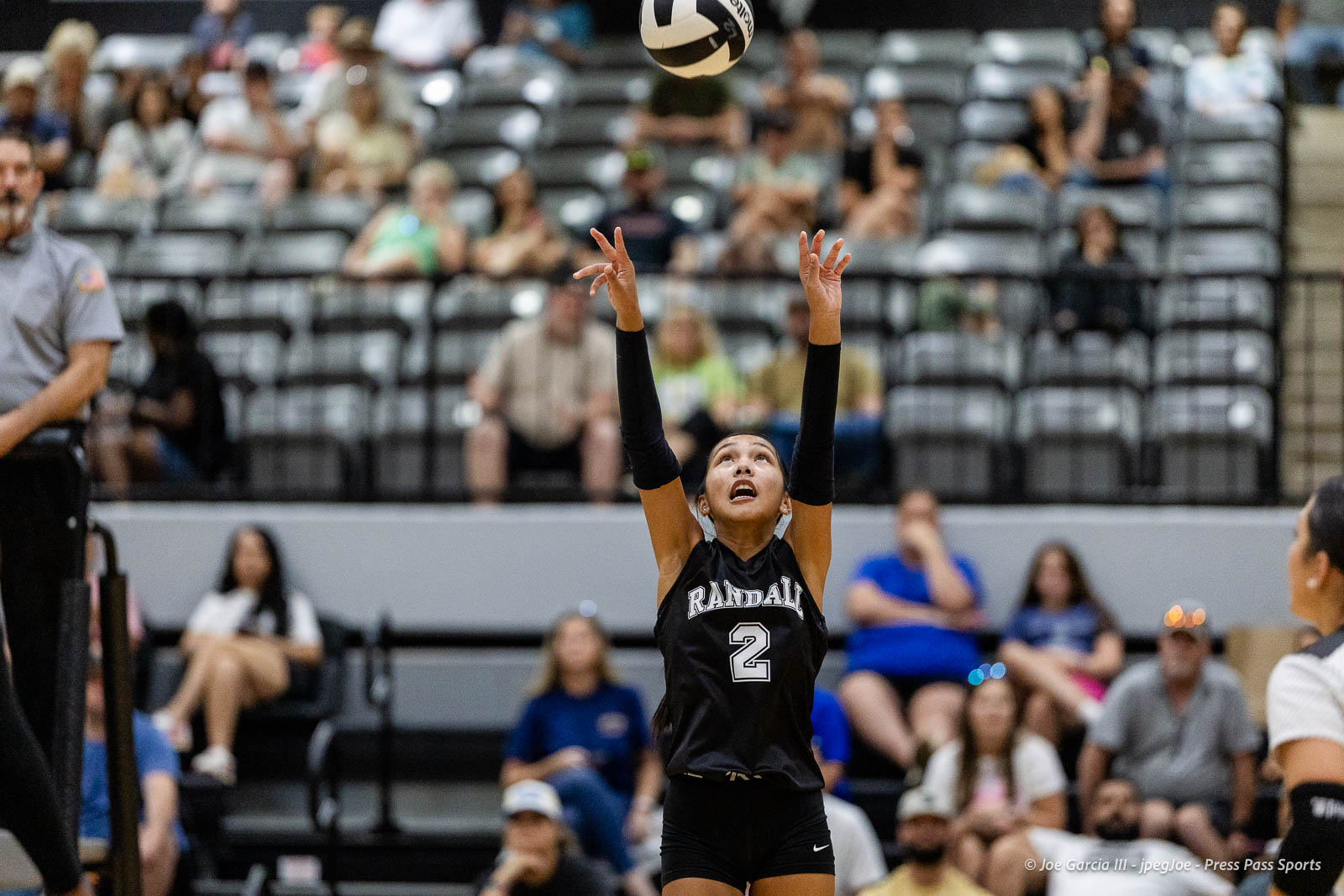 randall-showcase-2026-jpegJoe-pps-73 | Press Pass Sports Female volleyball player in a black Randall jersey #2 raises arms to set the ball above her in a gym with spectators in the stands a short distance away-to left is an official in gray.