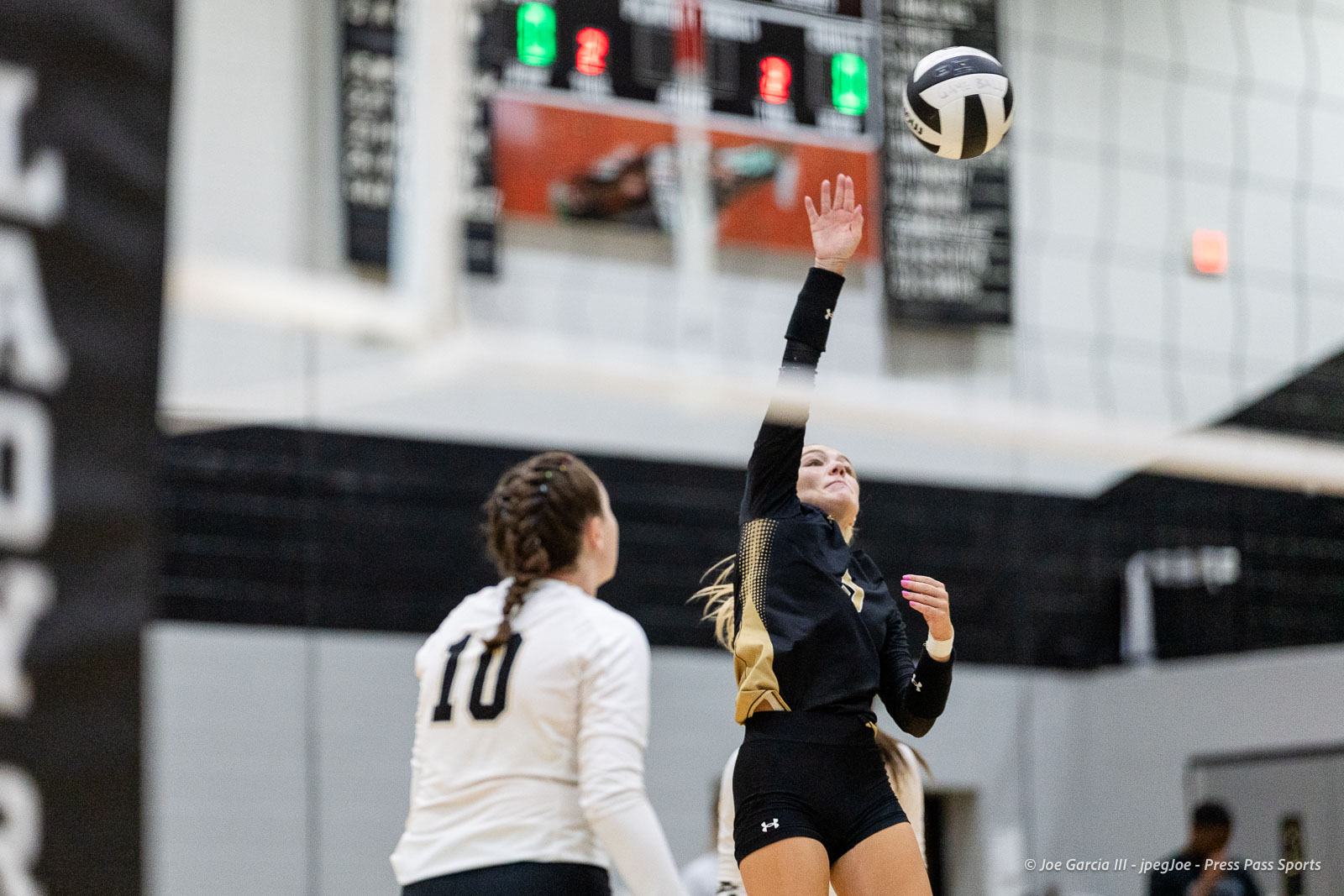 randall-showcase-2026-jpegJoe-pps-74 | Press Pass Sports Indoor volleyball game: a player in black reaches up to strike the ball while a teammate in white watches, scoreboard in the background.