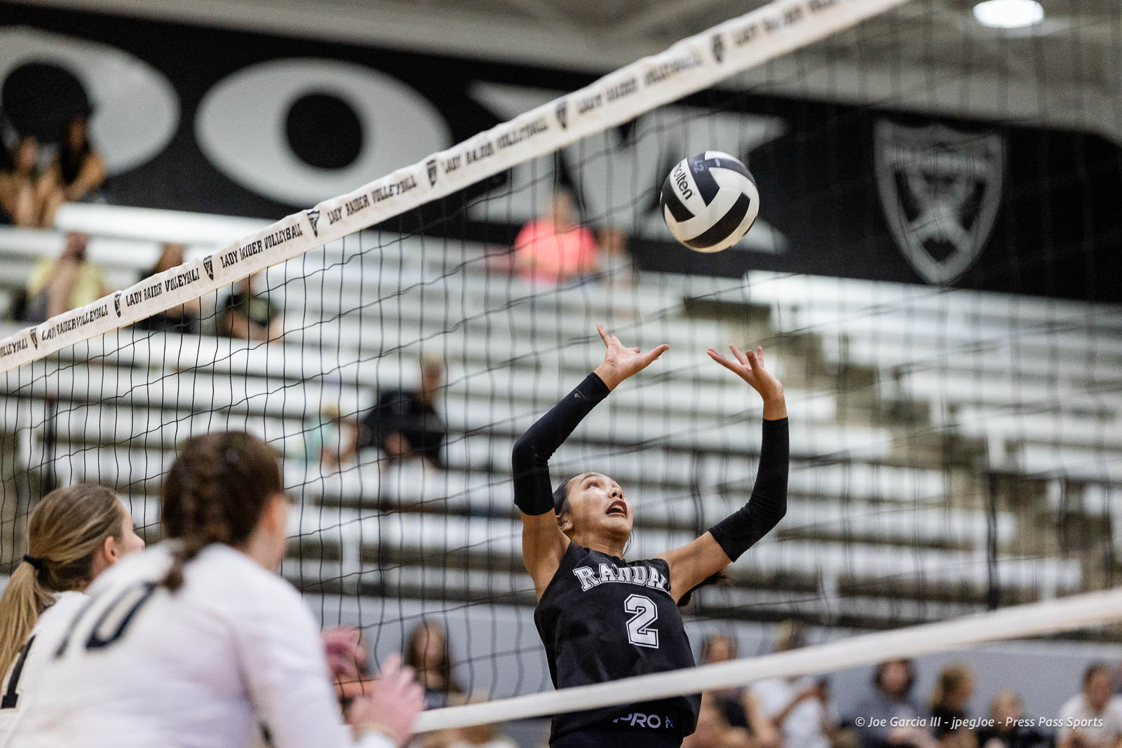 randall-showcase-2026-jpegJoe-pps-77 | Press Pass Sports Female volleyball player in a black jersey (#2) jumps near the net to set or spike the ball, spectators in bleachers behind her