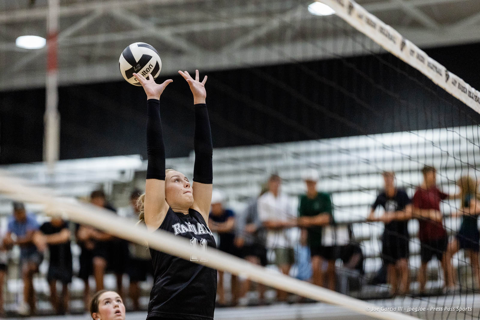 randall-showcase-2026-jpegJoe-pps-83 | Press Pass Sports Female volleyball player in a black uniform (#11) reaches upward to set the ball over the net with spectators blurred in the background.