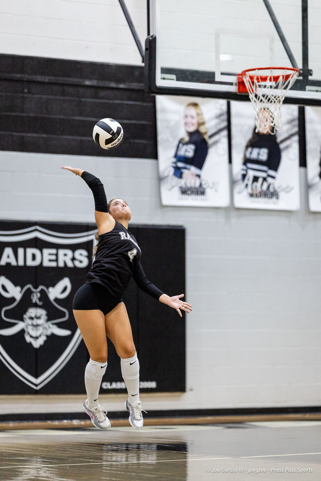 randall-showcase-2026-jpegJoe-pps-85 | Press Pass Sports Female volleyball player in black uniform serves the ball mid-air in a gym, Raiders banner in the background.