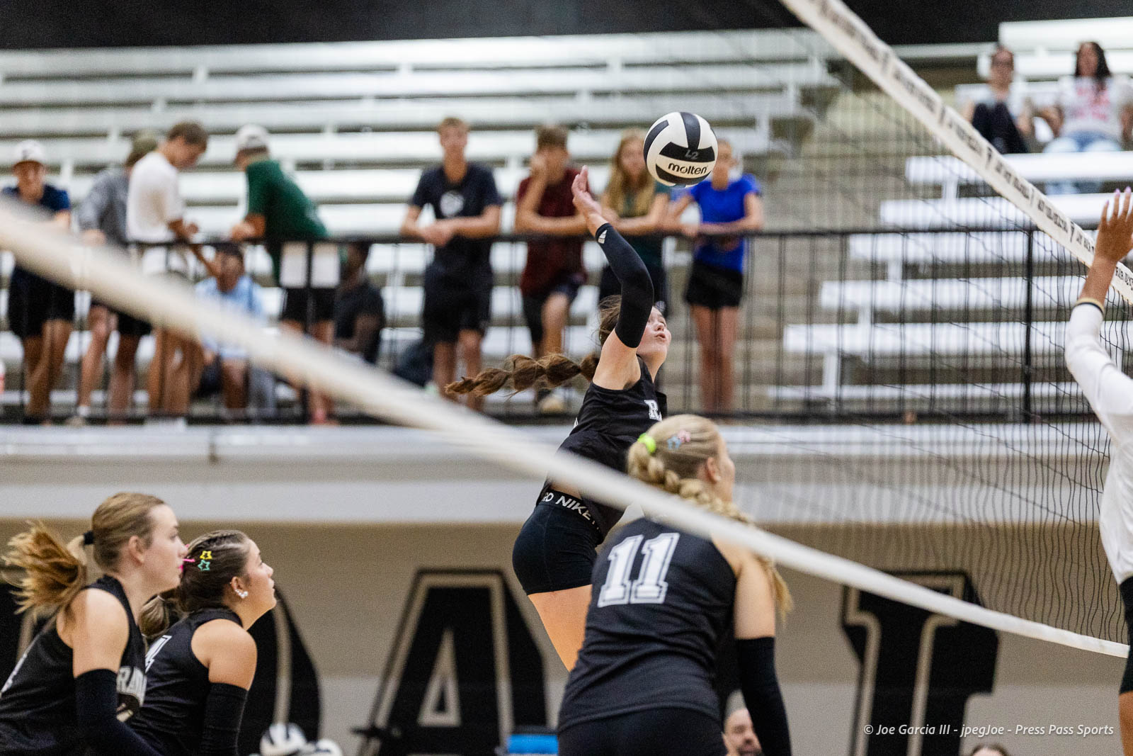 randall-showcase-2026-jpegJoe-pps-87 | Press Pass Sports Female volleyball player in a black jersey jumps to spike the ball over the net as teammates watch from the court.