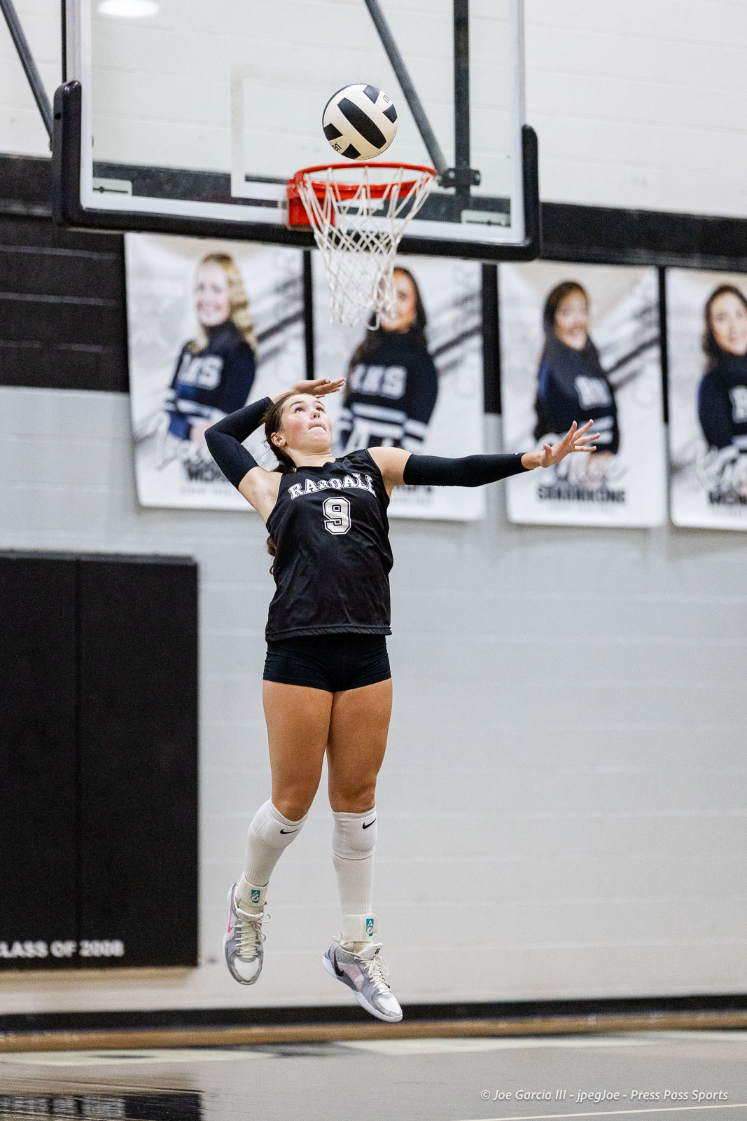 randall-showcase-2026-jpegJoe-pps-88 | Press Pass Sports Female volleyball player in a black jersey (#9) jumps in mid-air to spike a black-and-white ball during a match in a gym, with banners on the wall in the background.