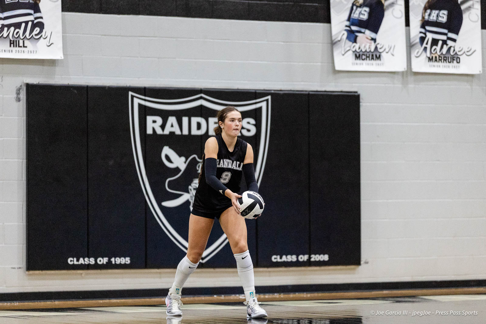 randall-showcase-2026-jpegJoe-pps-91 | Press Pass Sports Girl volleyball player in black uniform with jersey number 9, holding a ball on the court in front of a Raiders banner wall mural.