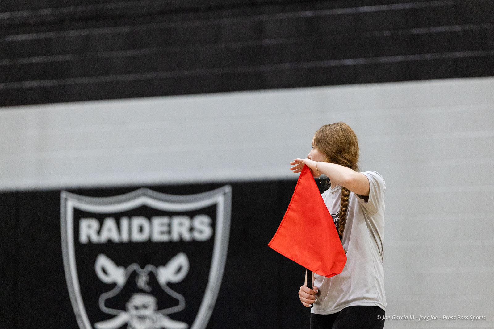 randall-showcase-2026-jpegJoe-pps-94 | Press Pass Sports Woman with a long braid wearing a light shirt holds an orange flag in a gym, Raiders banner visible in the background.