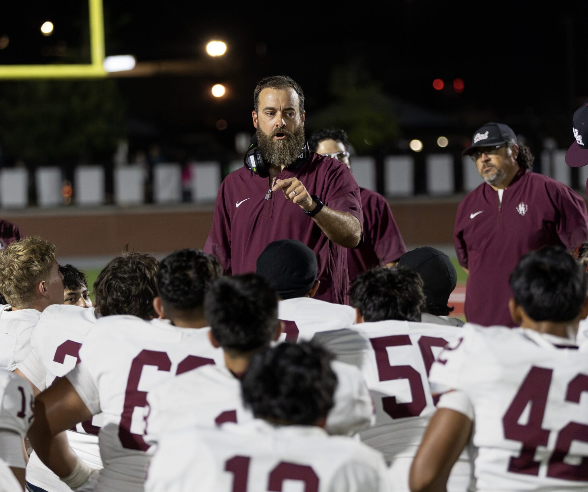 FW9A3069 | Press Pass Sports Bearded coach in maroon shirt and headset directing a night-time football huddle with players in white jerseys gathered around him.