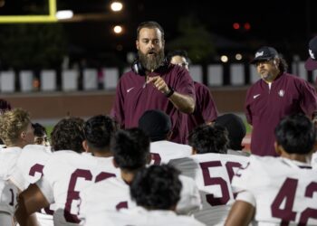 FW9A3069 | Press Pass Sports Bearded coach in maroon shirt and headset directing a night-time football huddle with players in white jerseys gathered around him.