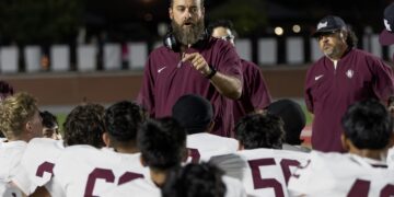 Bearded coach in maroon shirt and headset directing a night-time football huddle with players in white jerseys gathered around him.