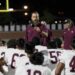 FW9A3069 | Press Pass Sports Bearded coach in maroon shirt and headset directing a night-time football huddle with players in white jerseys gathered around him.