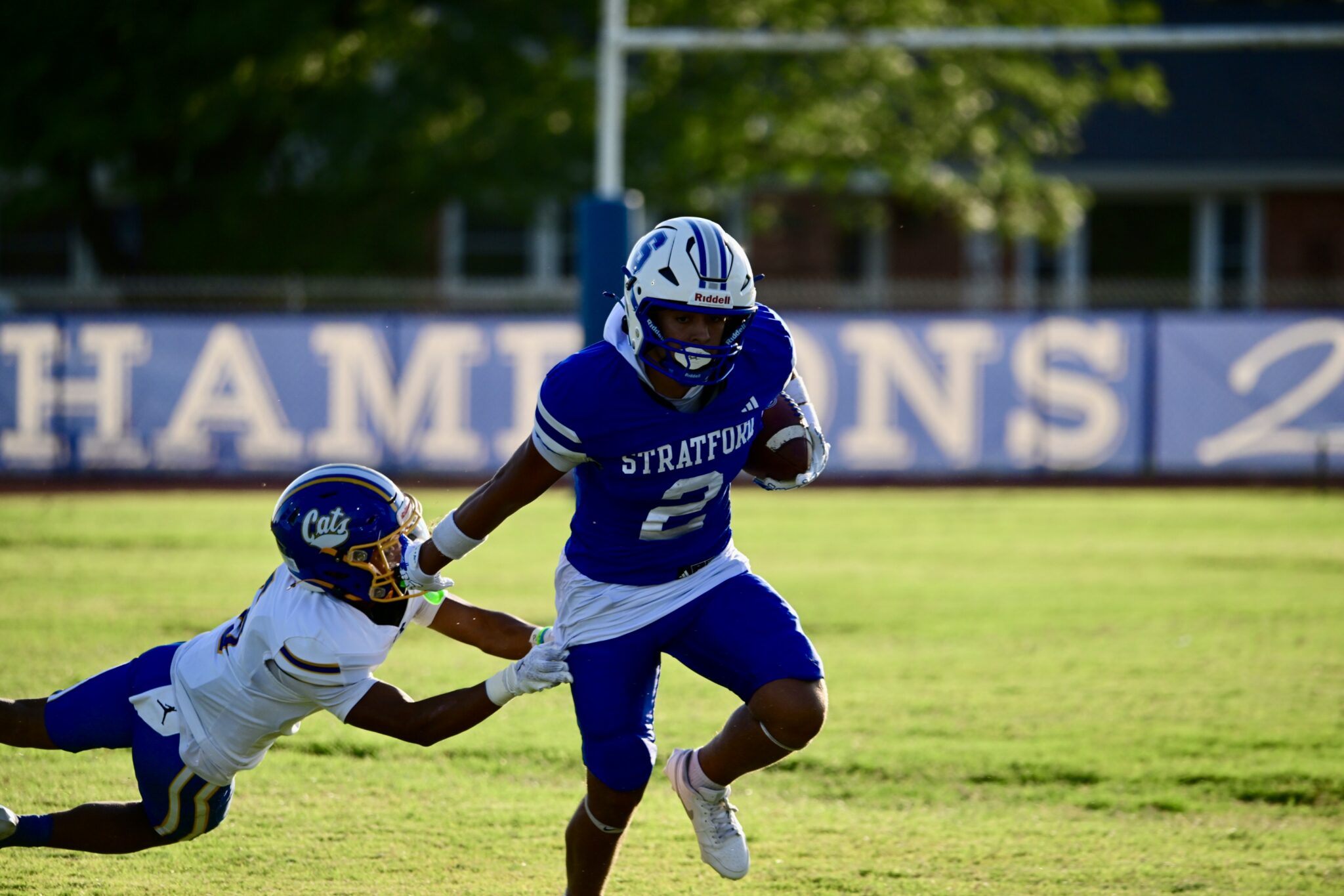 image1 25 | Press Pass Sports Two lacrosse players in blue and white uniforms race on a sunny field, one in front with a helmet and mouthguard while the other dives from the side ahead of him.