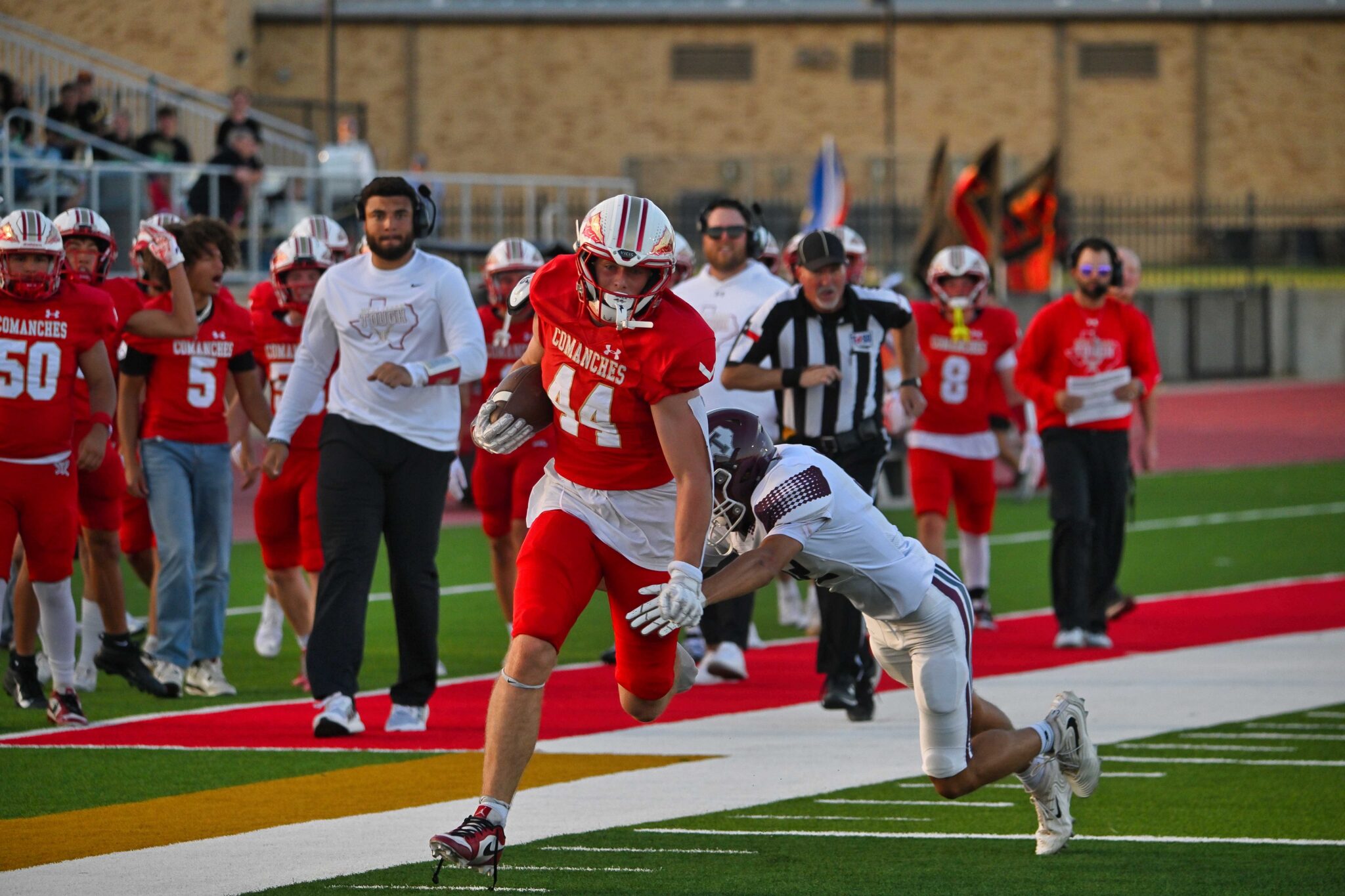 Red-team football player (No. 44) in a sprint with the ball, pursued by a defender on a green artificial turf field as teammates and crowd look on.