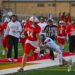 image6 7 | Press Pass Sports Red-team football player (No. 44) in a sprint with the ball, pursued by a defender on a green artificial turf field as teammates and crowd look on.
