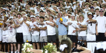 Large group of students in white shirts cheering with arms raised at a stadium crowd.